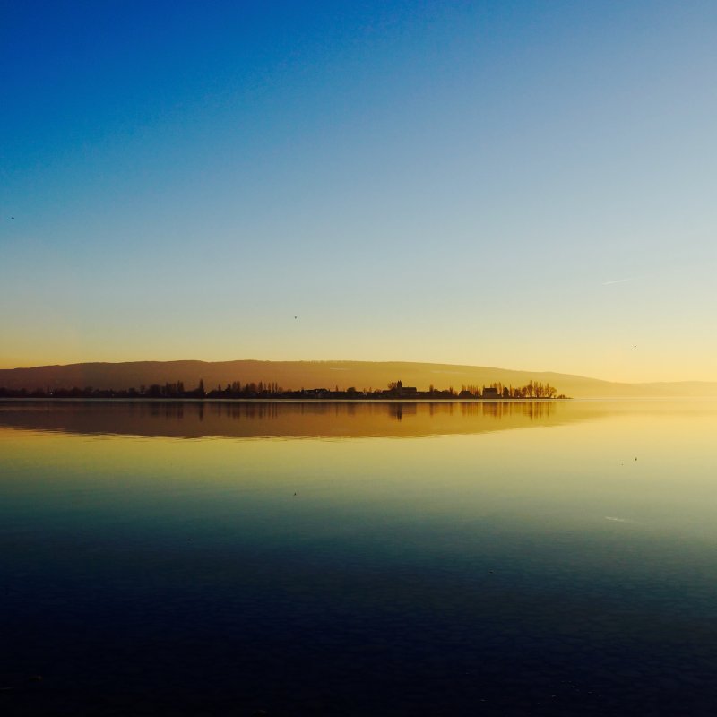 Ruhiger See bei Sonnenuntergang. Landschaft mit Bergen, Bäumen und blauem Himmel. Spiegelung im Wasser.