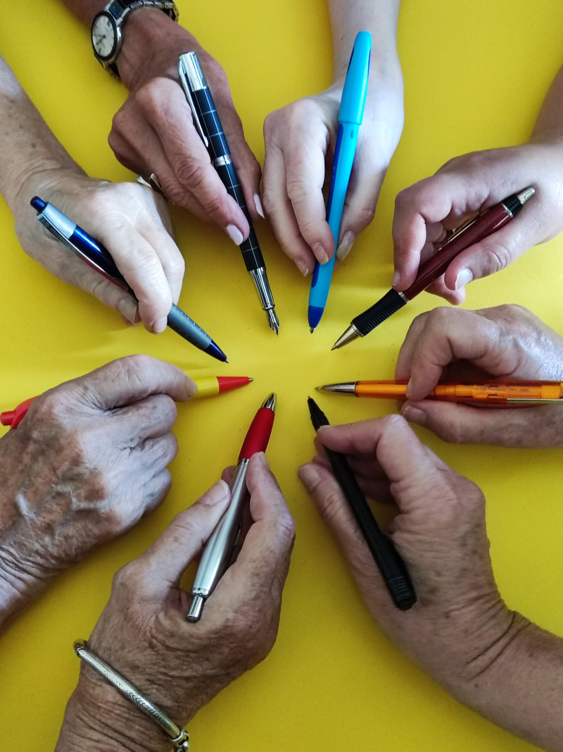Hände halten Stifte. Schreibgeräte in verschiedenen Farben auf gelbem Grund. Kugelschreiber, Füller.