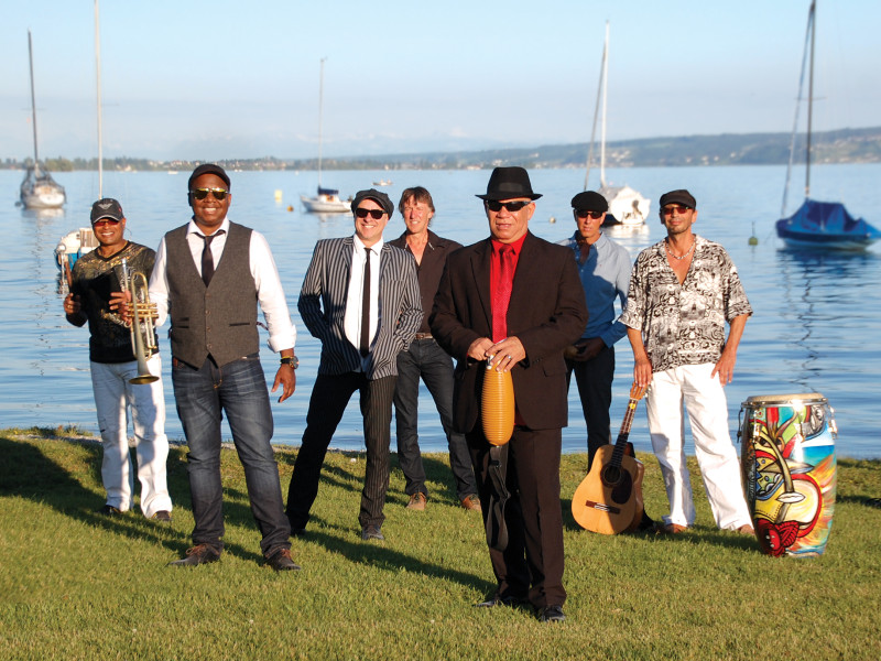 Cuban band performing outdoors. Musicians with instruments: trumpet, guitar, conga. Lake and sailboats in background.