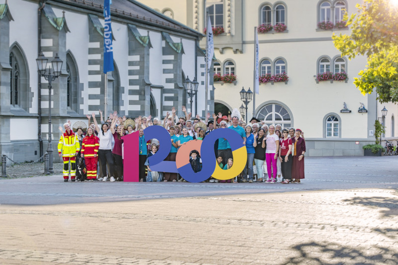 Gruppenfoto mit "1200" Zahl vor historischer Architektur. Menschen feiern Jubiläum. Vielfalt und Gemeinschaft im Stadtbild.