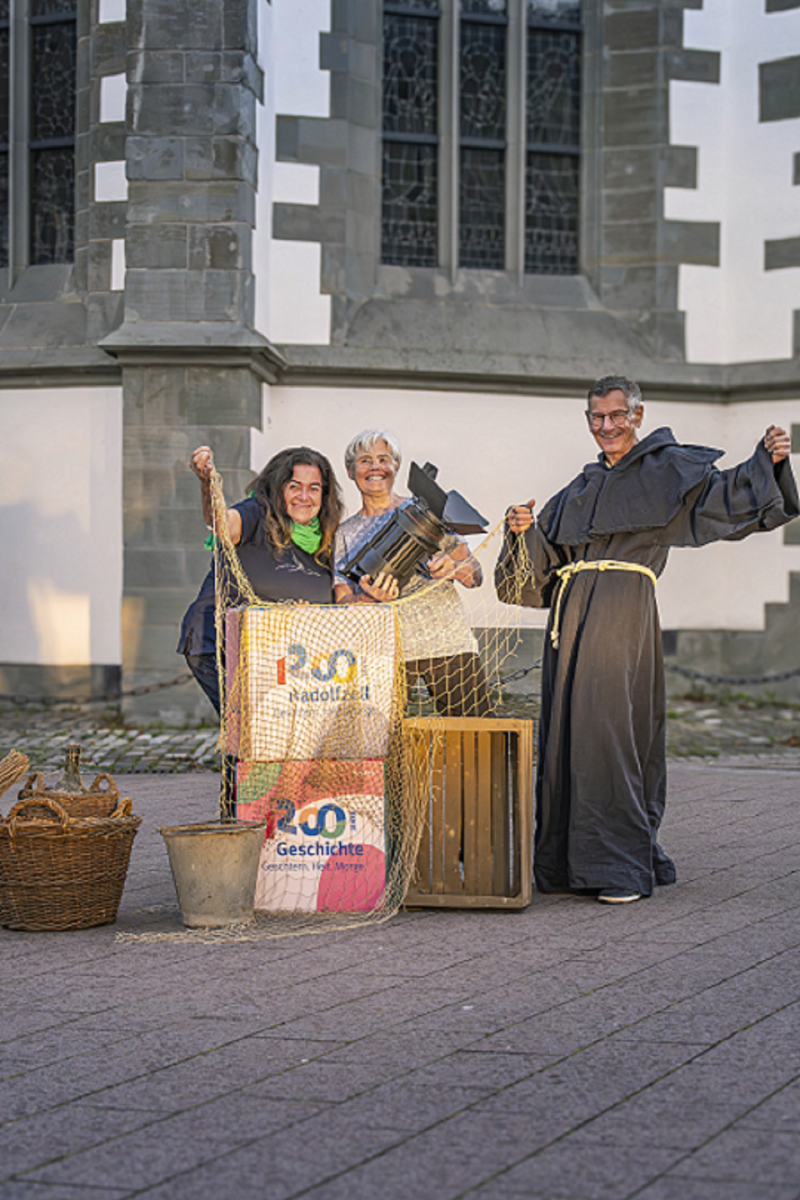 Drei Personen vor Kirche mit Requisite: Scheinwerfer, Fischernetz, Körbe, Kisten. Radolfzell Geschichte.