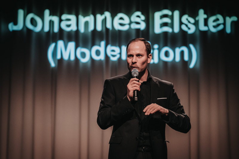 Johannes Elster, Moderator auf der Bühne mit Mikrofon, im schwarzen Anzug. Bühnenbeleuchtung im Hintergrund.