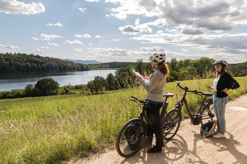 Zwei Radfahrerinnen mit Helmen und Fahrrädern auf einem Feldweg mit Blick auf einen See und bewaldete Hügel.