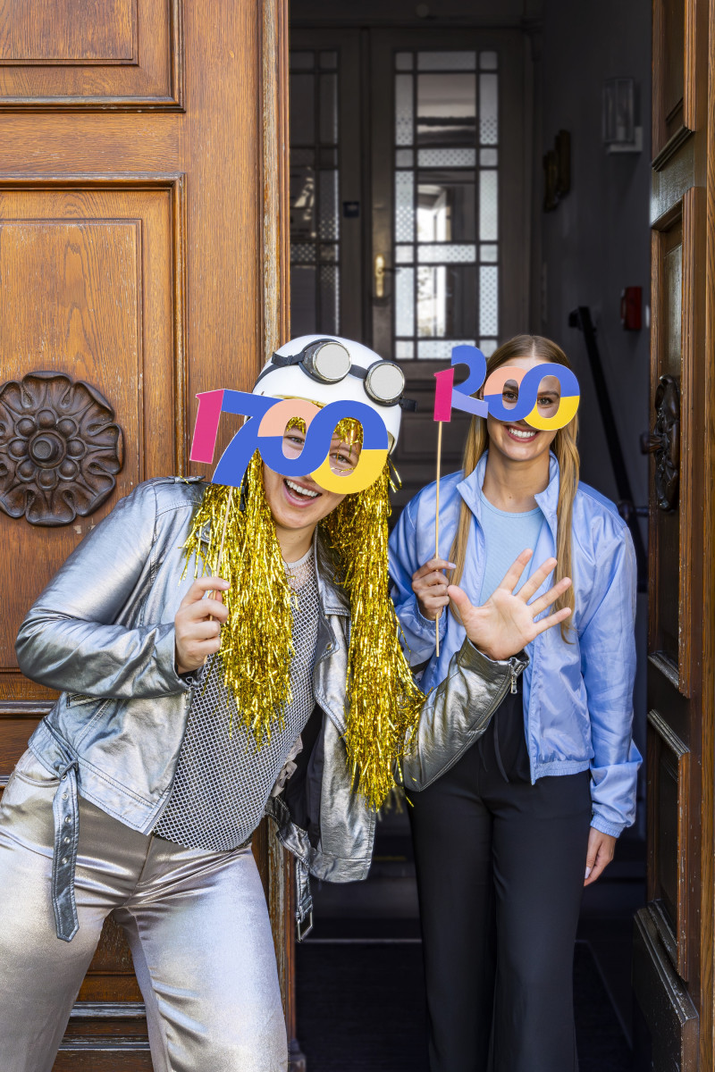Zwei Frauen feiern mit 700 Masken vor einer Tür, festliche Kleidung und goldene Lametta-Perücken, fröhliche Stimmung.